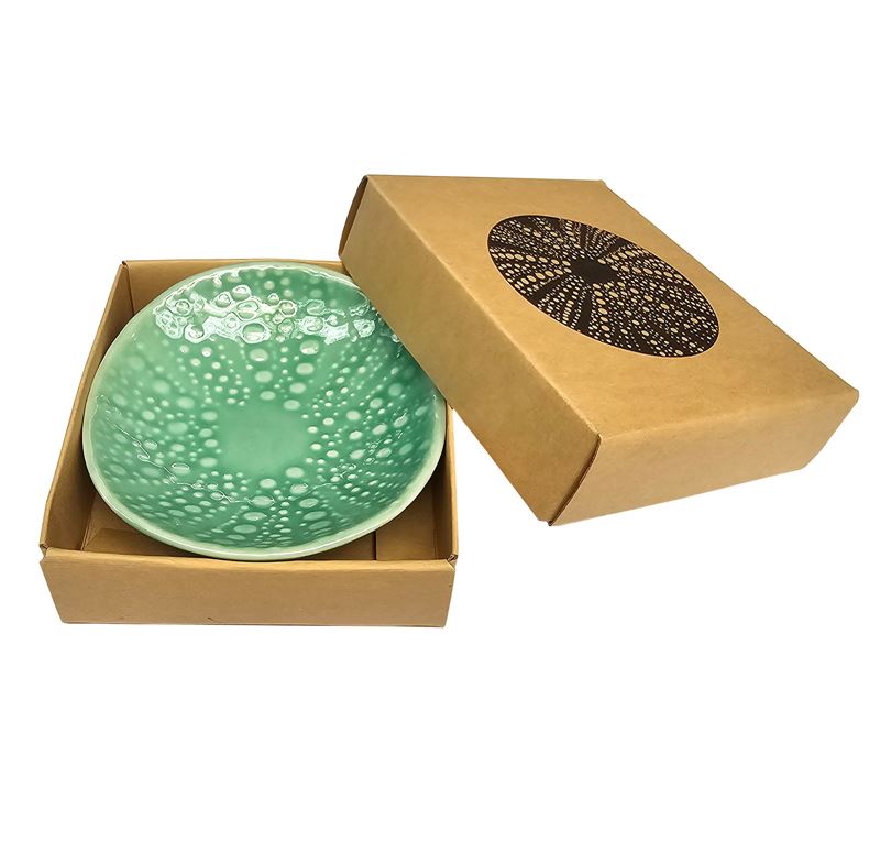 A green porcelain bowl with a textured design, presented in an open cardboard box with a lid featuring a round black pattern.