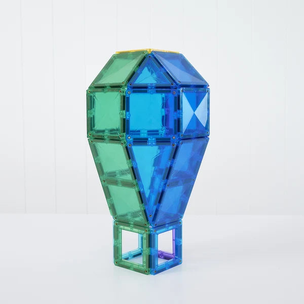 A geometric, faceted toy structure built from translucent magnetic building tiles, standing upright on a white surface against a white paneled background. The structure features shades of blue, green, and teal.