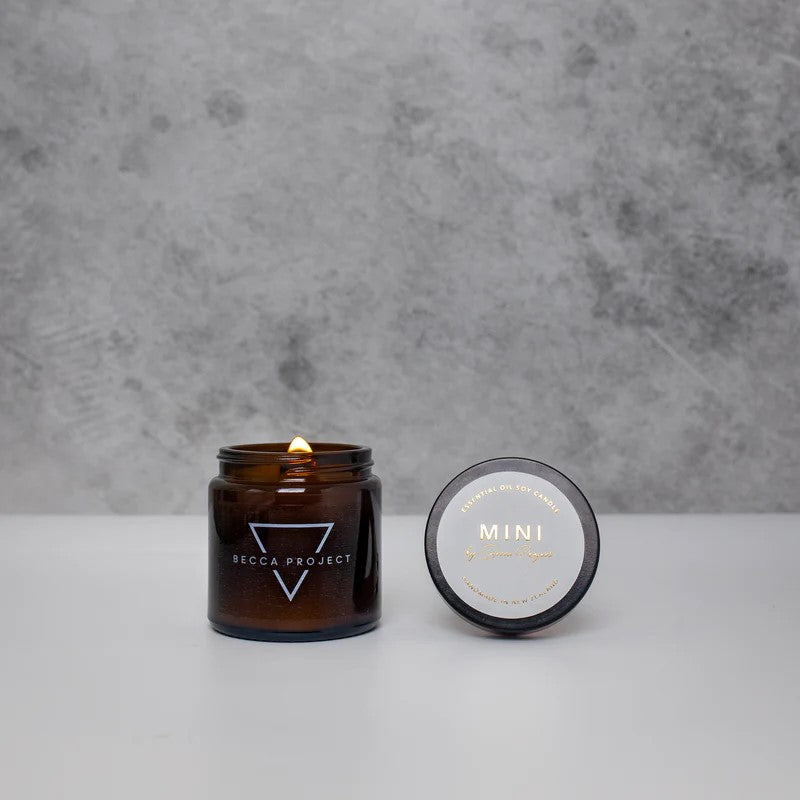 A small amber glass candle jar with a lit candle inside, alongside its lid that has 'MINI' printed on it