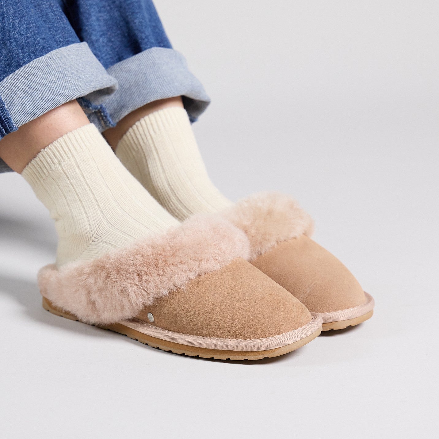 Slip on Sheepskin Slippers on a ladies feet with cream coloured socks below denim jeans on a white background