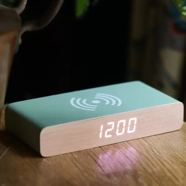 A rectangular digital clock with a light wood grain front and a mint green top surface featuring a white wireless charging symbol. The clock displays '12:00' on its front face and sits on a wooden table next to a plant.