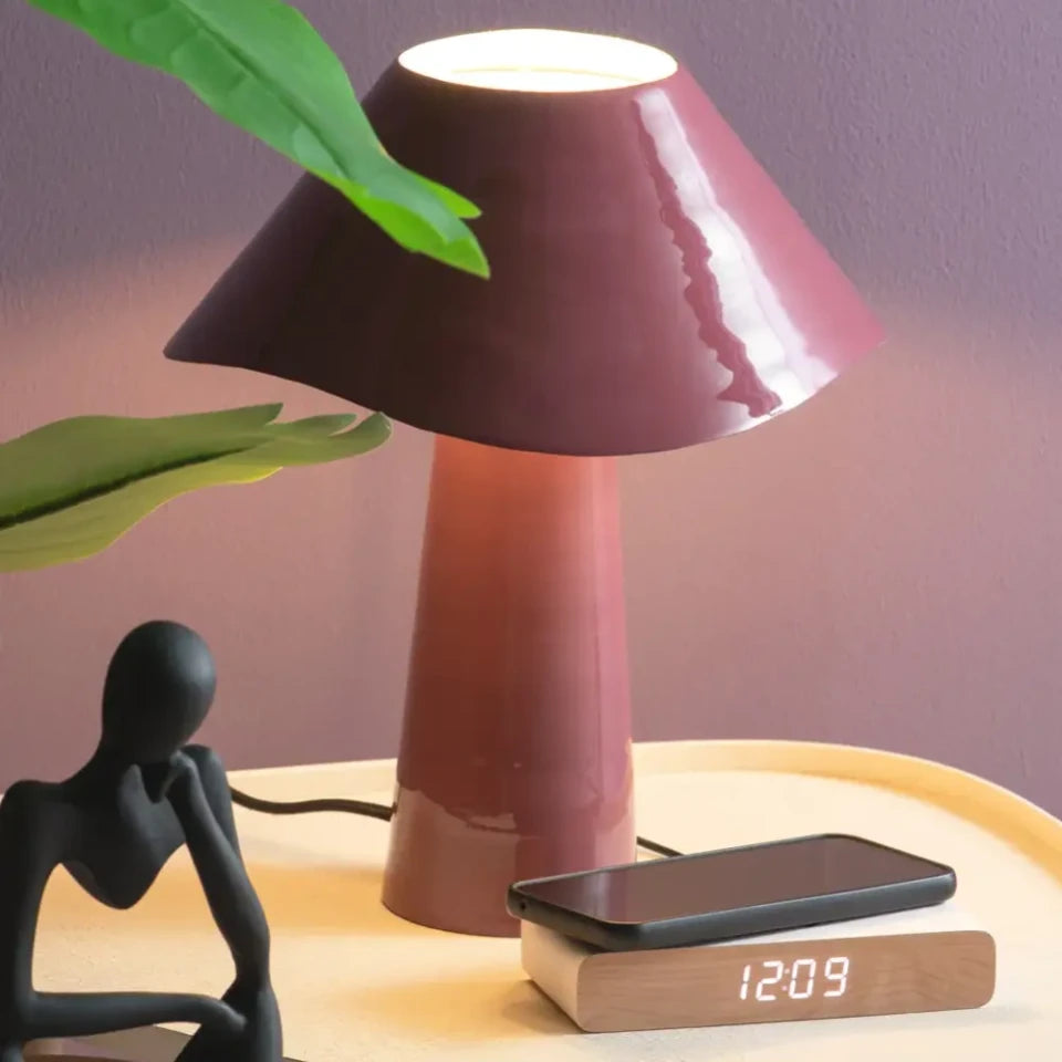 A mushroom-shaped table lamp with a glossy burgundy shade and matching stem, glowing softly on a light wood nightstand. A smartphone rests on top of the lamp, and a small black figurine and green leaves are visible in the scene.