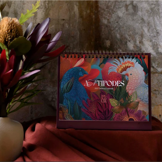 A spiral-bound 2023 Tiodes calendar featuring colorful tropical bird illustrations, styled on draped rust-red fabric next to a vase with flowers against a textured stone wall.