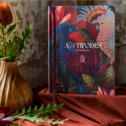 ANTIPODES 2027 Weekly Diary standing upright on draped rust-colored fabric, surrounded by a wooden vase with dried flowers and green leaves. The cover features a highly detailed, colorful illustration of a blue bird, a pink-and-white cockatoo, and tropical flowers.