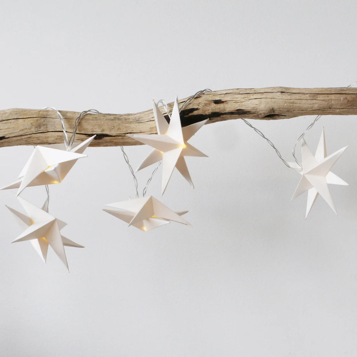 White paper star string lights glowing with warm yellow light, draped over a rustic wooden branch