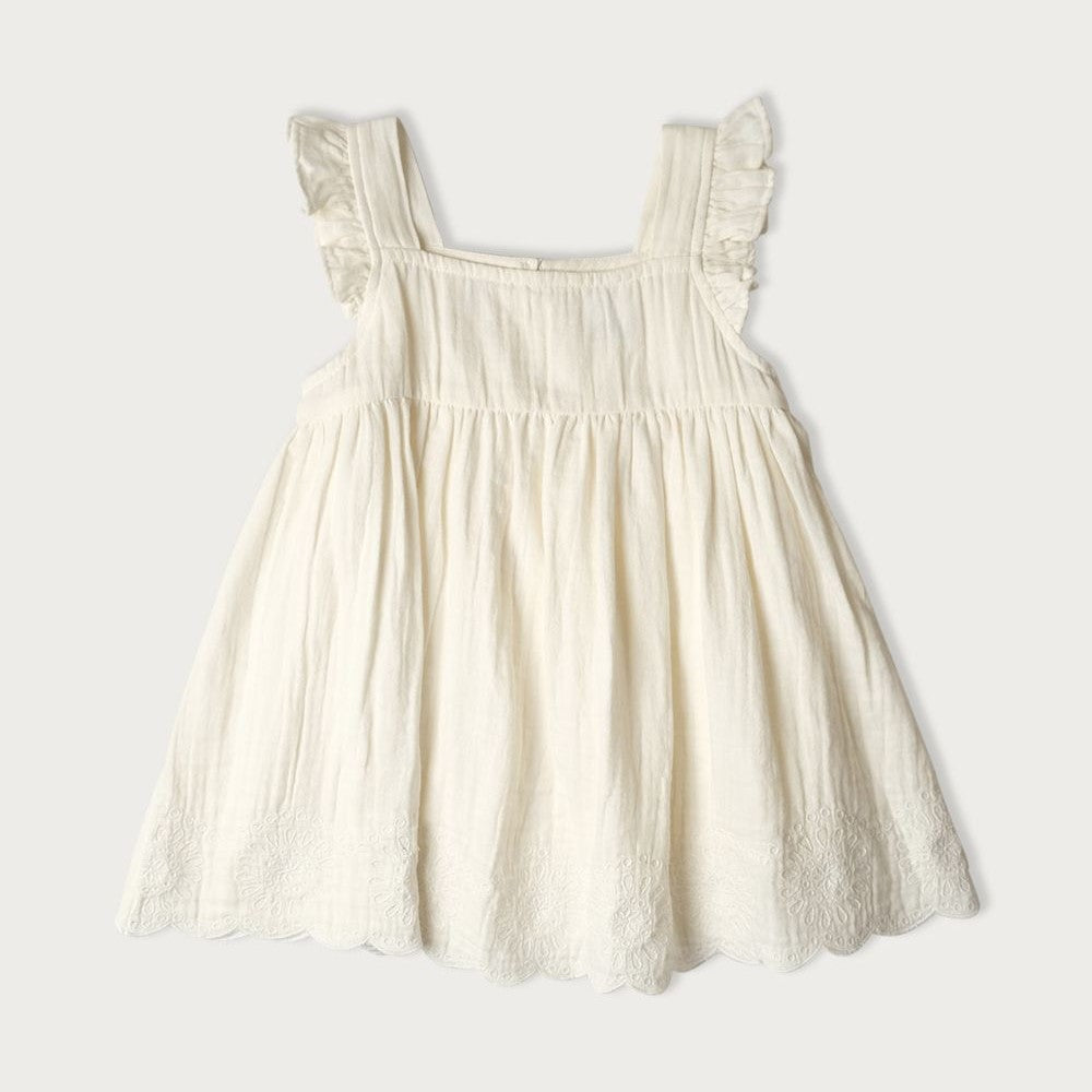 A sleeveless, vanilla-colored muslin dress with embroidery, scalloping, and ruffle detailing, featuring a wooden button closure at the back.