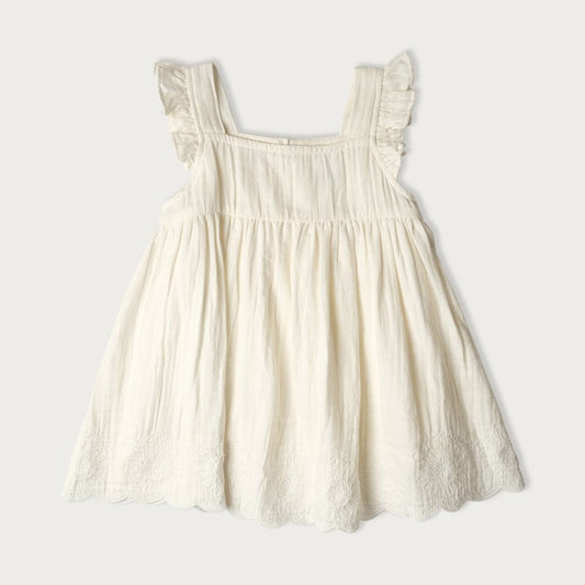 A sleeveless, vanilla-colored muslin dress with embroidery, scalloping, and ruffle detailing, featuring a wooden button closure at the back.