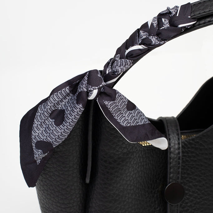 A close-up of a black pebbled leather handbag with a black and white patterned silk scarf tied to its handle,
