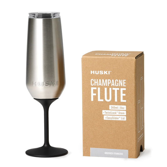 A stainless steel Huski Champagne Flute with a wide mouth and a black base, displayed next to its packaging box.
