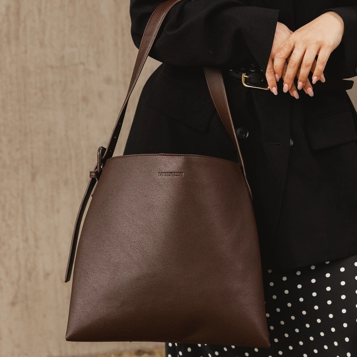 Brown vegan leather handbag held by a person wearing a black coat and polka dot skirt.