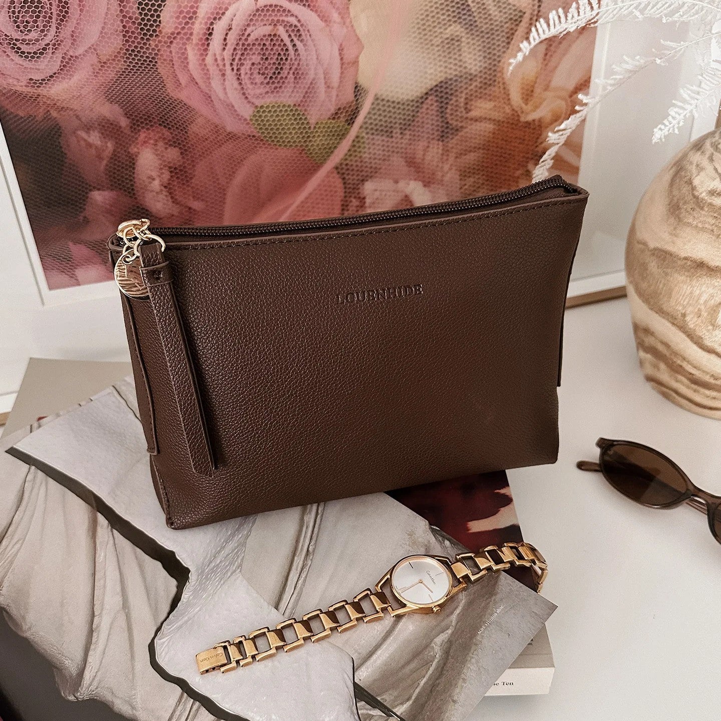 Brown vegan leather clutch bag with a gold watch on a white surface with flowers in the background