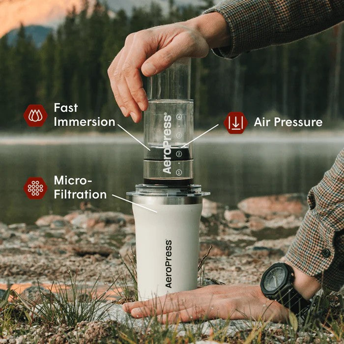 Person using an Aeropress coffee maker outdoors by a lake