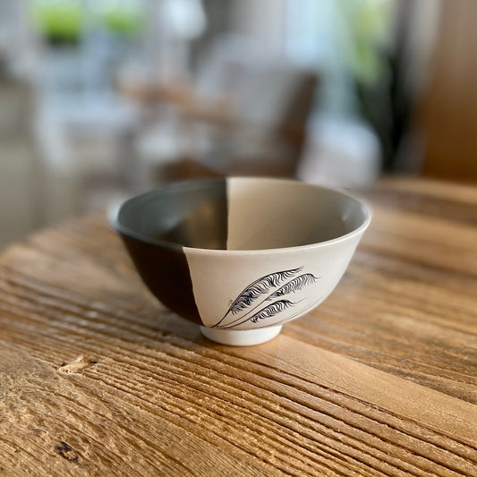 A white porcelain bowl with a black and white coastal toetoe design, placed on a wooden table.