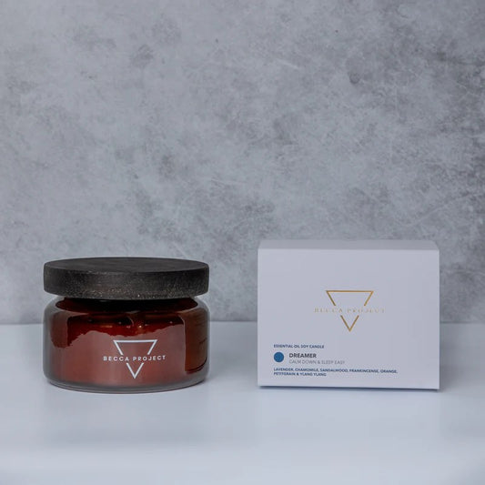 A medium-sized Dreamer candle in a brown jar with a black lid, placed next to its white packaging box with the Becca Project branding.