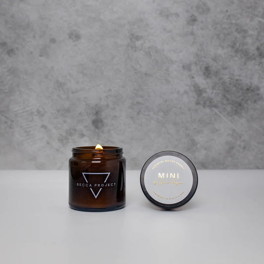A small amber glass candle jar with a lit candle inside, alongside its lid that has 'MINI' printed on it