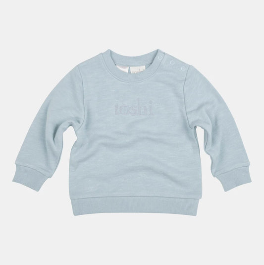 A blue organic cotton sweater for babies, with long sleeves and the Toshi brand logo embroidered on the front.