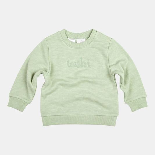 A green organic cotton sweater for babies with the Toshi logo embroidered on it.