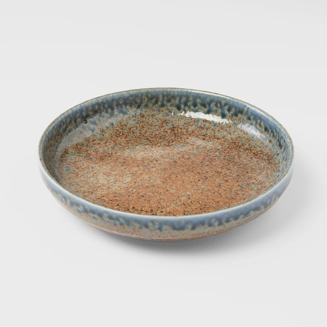 A shallow round ceramic plate with a speckled glaze, featuring a warm brown center and a blue-gray rim, photographed on a light gray background.