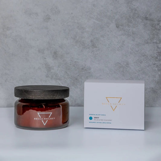 A medium-sized 'ENVY' candle by Becca Project, with a dark lid and a white label, placed next to its packaging box.