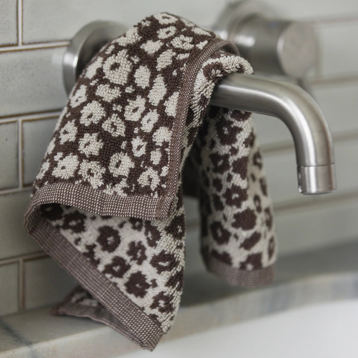 Brown and white patterned towel hanging on a silver faucet against a tiled wall.