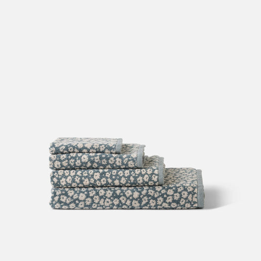 A stack of four folded bathroom set pieces, likely a towel set, featuring a small white floral pattern on a muted blue-gray background, set against a plain light gray studio background.