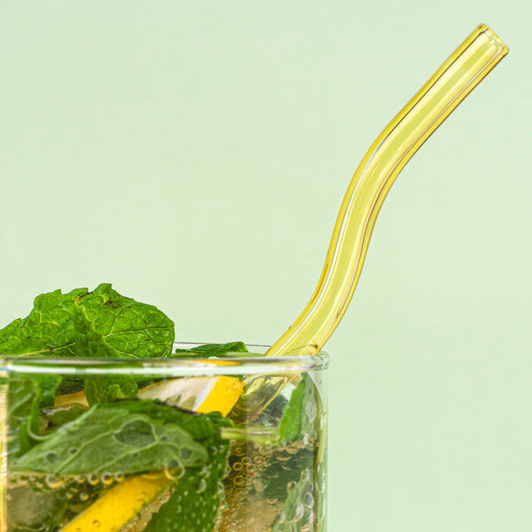 A clear glass filled with a lemon and mint beverage, featuring fresh mint leaves, lemon slices, and a curved translucent yellow straw, set against a pale mint green background.
