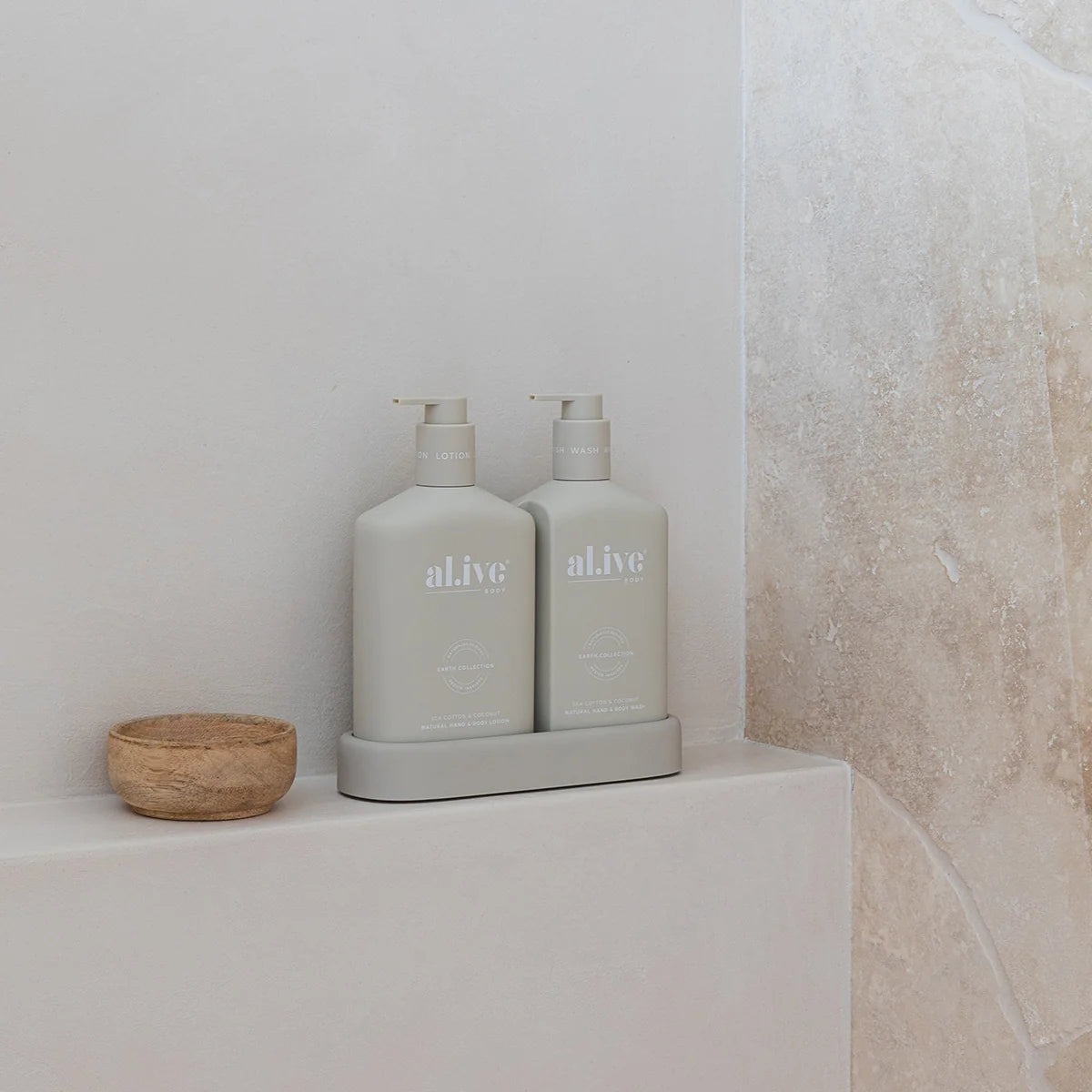 Two light gray pump bottles labeled 'alive' on a matching base, placed on a white ledge beside a small wooden bowl, with a textured beige wall in the background.
