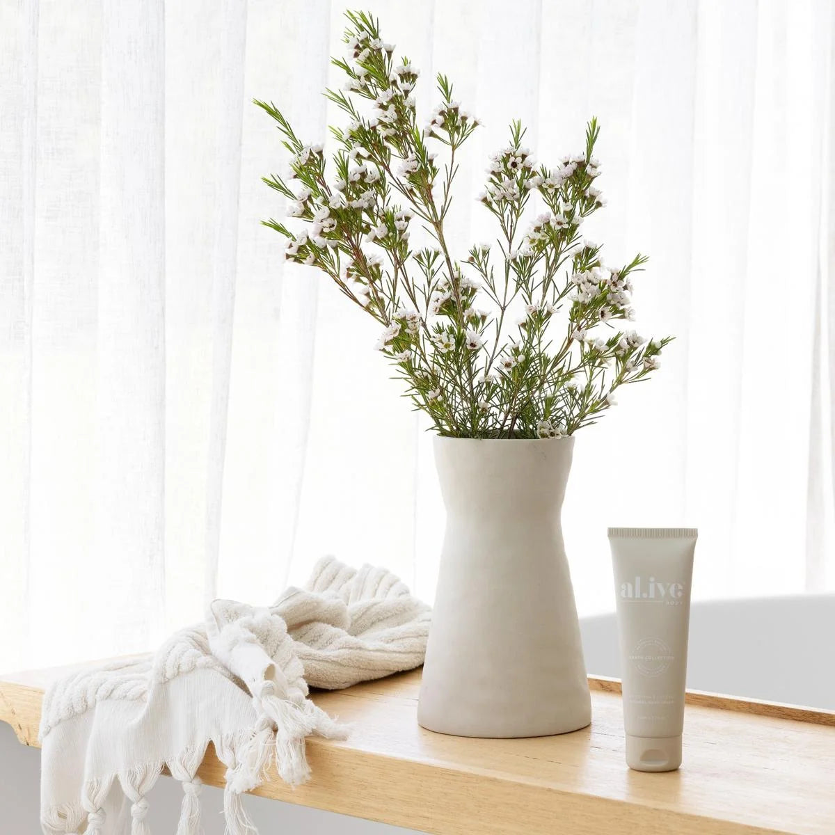 A matte off-white ceramic vase with a wavy, flared shape holding green sprigs, styled on a light wood shelf next to a folded cream knit blanket and a white tube labeled 'alive' in front of a soft white curtain background.