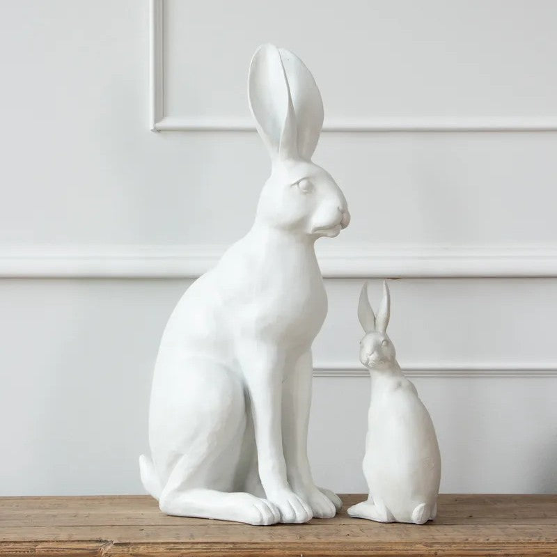 A pair of white rabbit figurines, one large seated rabbit and one small seated rabbit, displayed on a wooden surface against a white paneled wall.