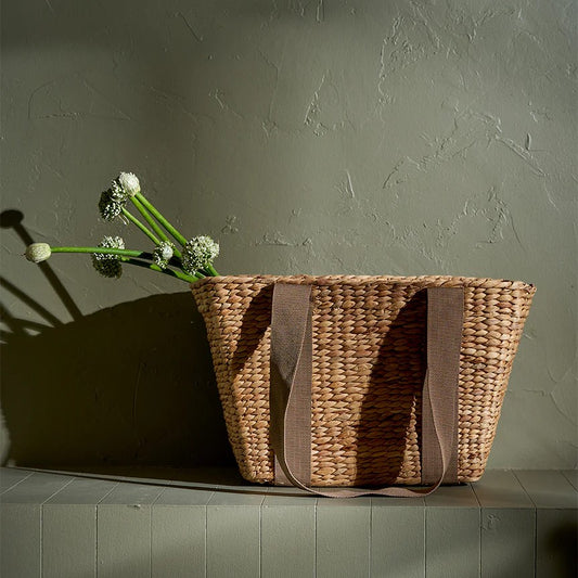 A wicker tote bag with brown canvas handles resting on a surface, with a bunch of flowers partially visible inside it