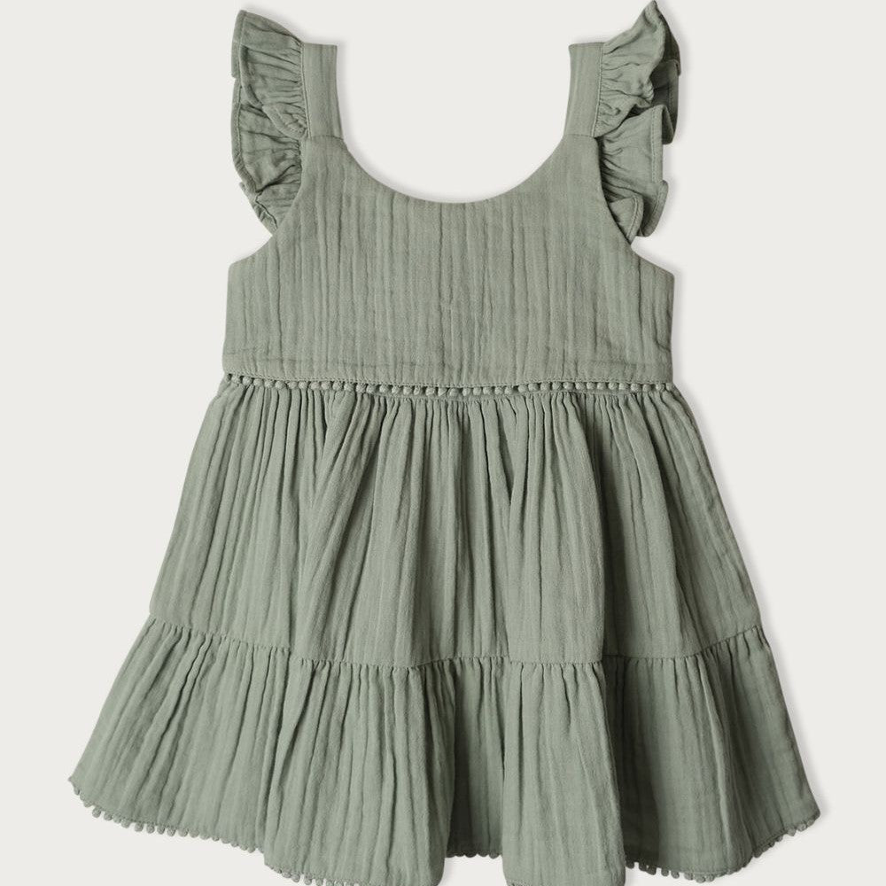 A short-sleeved, green, muslin baby dress with embroidered pompoms and ruffles on the shoulders, featuring a gathered bottom tier.
