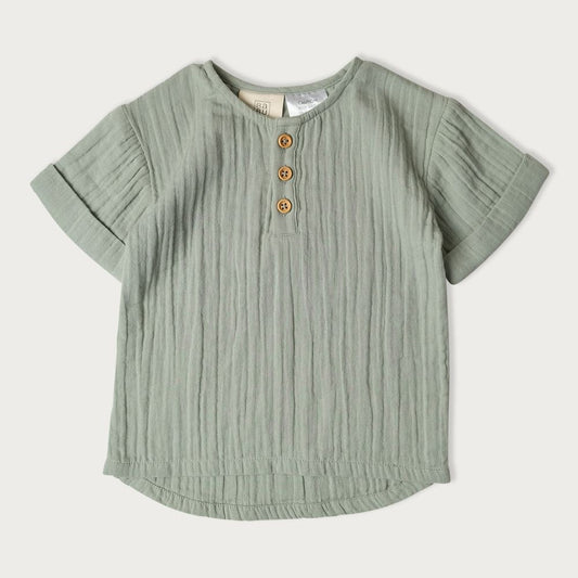 A light green, short-sleeved baby shirt with wooden buttons in a row down the center, featuring a relaxed grandpa style fit with a rounded hem.