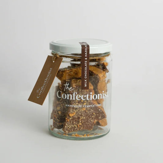 A glass jar filled with hazelnut toffee pieces, covered in milk chocolate and sprinkled with crushed hazelnuts, with a label that reads 'The Confectionists'.