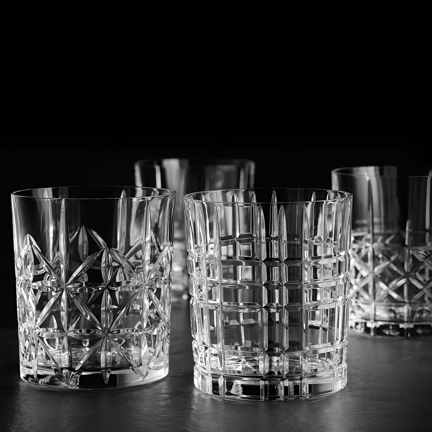 A set of clear cut-crystal whiskey glasses with geometric patterns, arranged on a dark surface against a black background.
