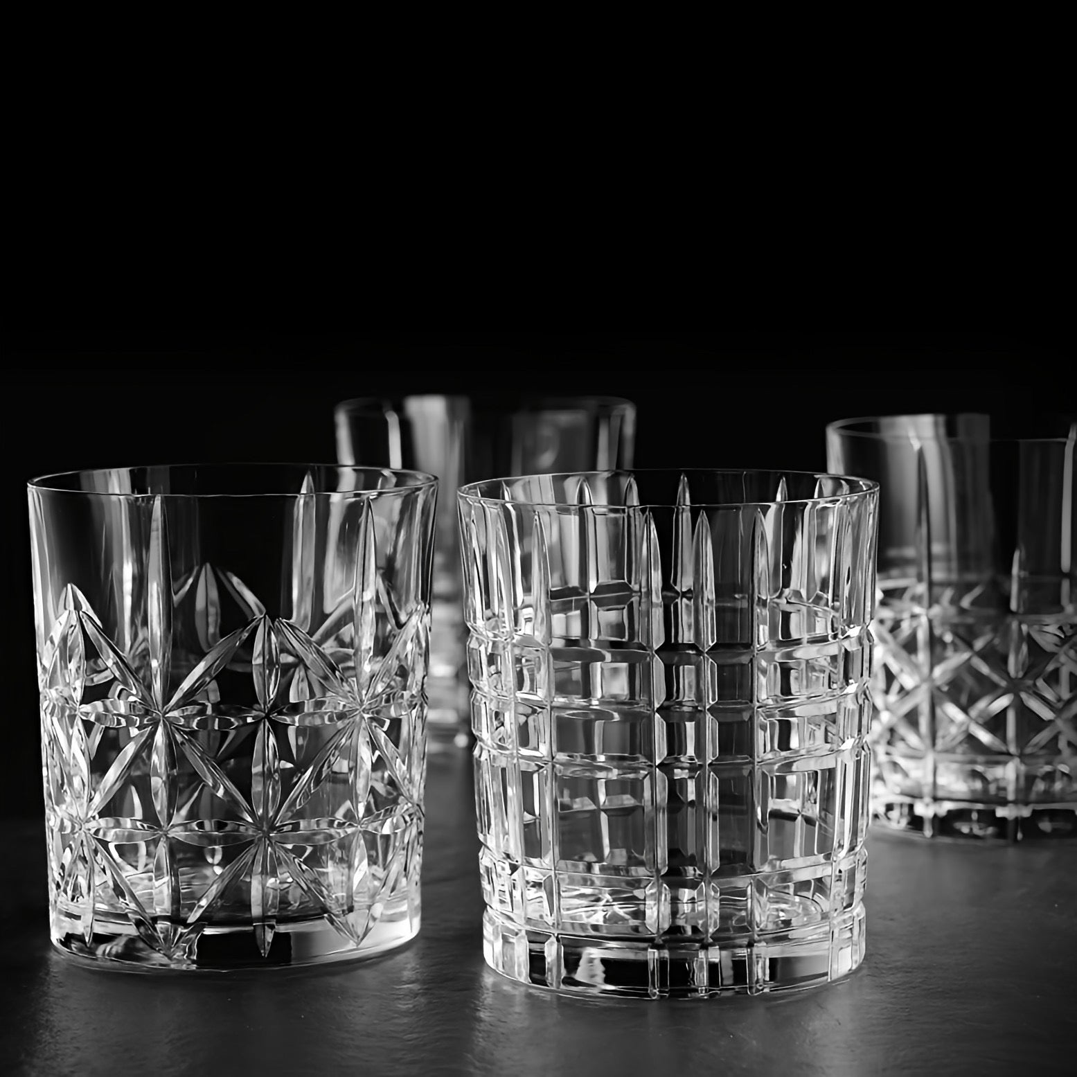 A set of clear cut-crystal whiskey glasses with geometric patterns, arranged on a dark surface against a black background.
