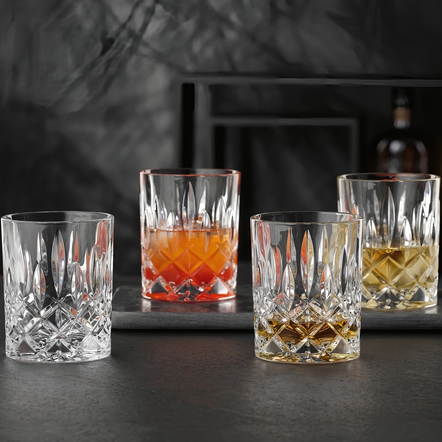 Four clear cut-crystal whiskey glasses arranged on a dark tray against a moody dark gray background. Three glasses are empty, and one is filled with amber liquid.