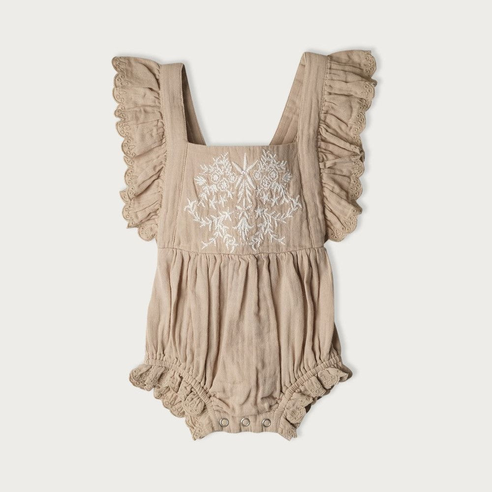 A beige-colored baby bodysuit with embroidered detailing on the front and ruffle trim, featuring an elastic waistband and adjustable tie at the back.