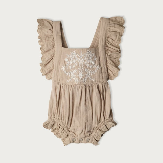 A beige-colored baby bodysuit with embroidered detailing on the front and ruffle trim, featuring an elastic waistband and adjustable tie at the back.