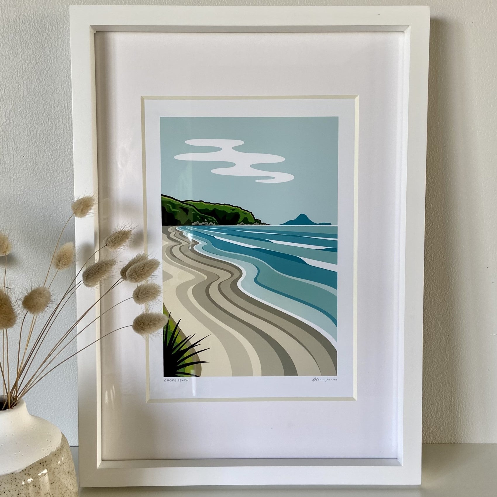 A framed print featuring a coastal scene with a beach, sea, and hills, Ohope A3 size, displayed on a white wall.