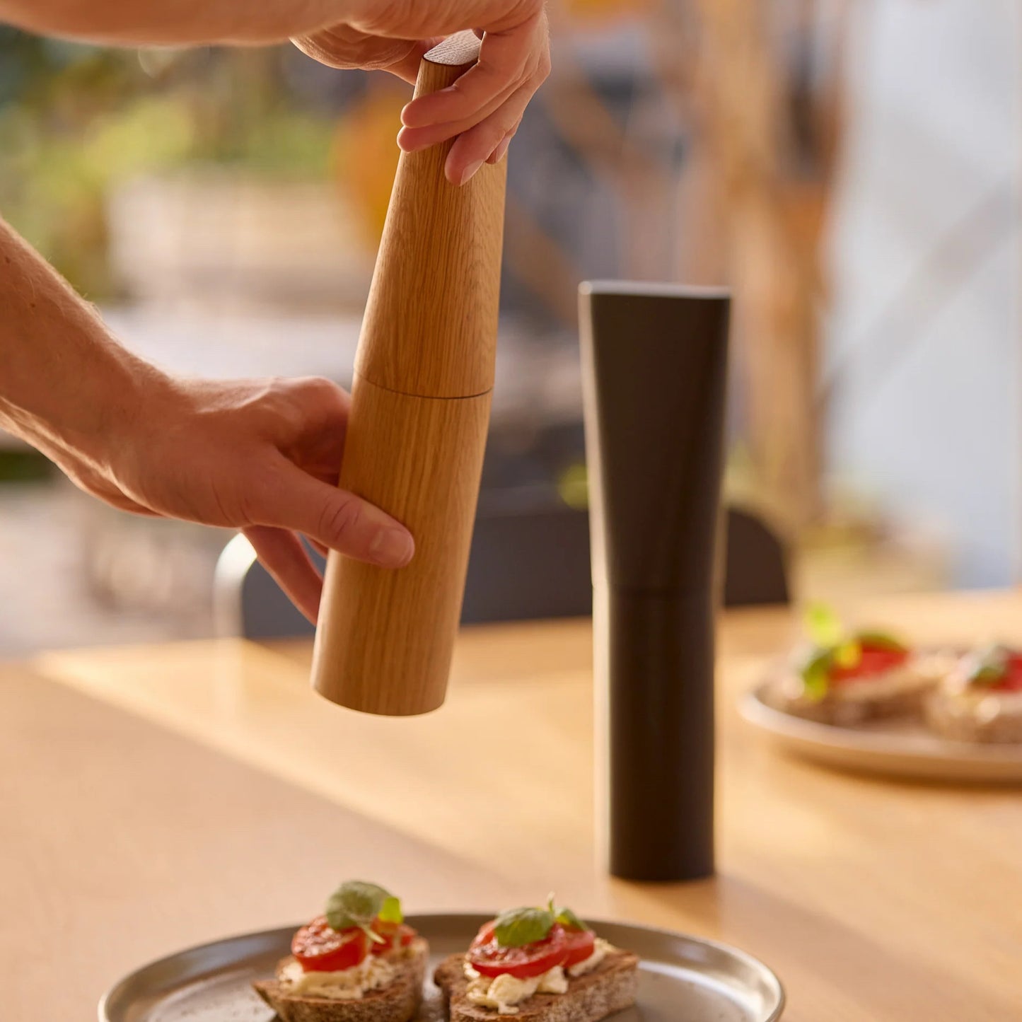 A person's hands are shown using a wooden pepper mill to grind pepper over small toast on a table, with a dark cylindrical container standing nearby in a warm, softly blurred kitchen setting.