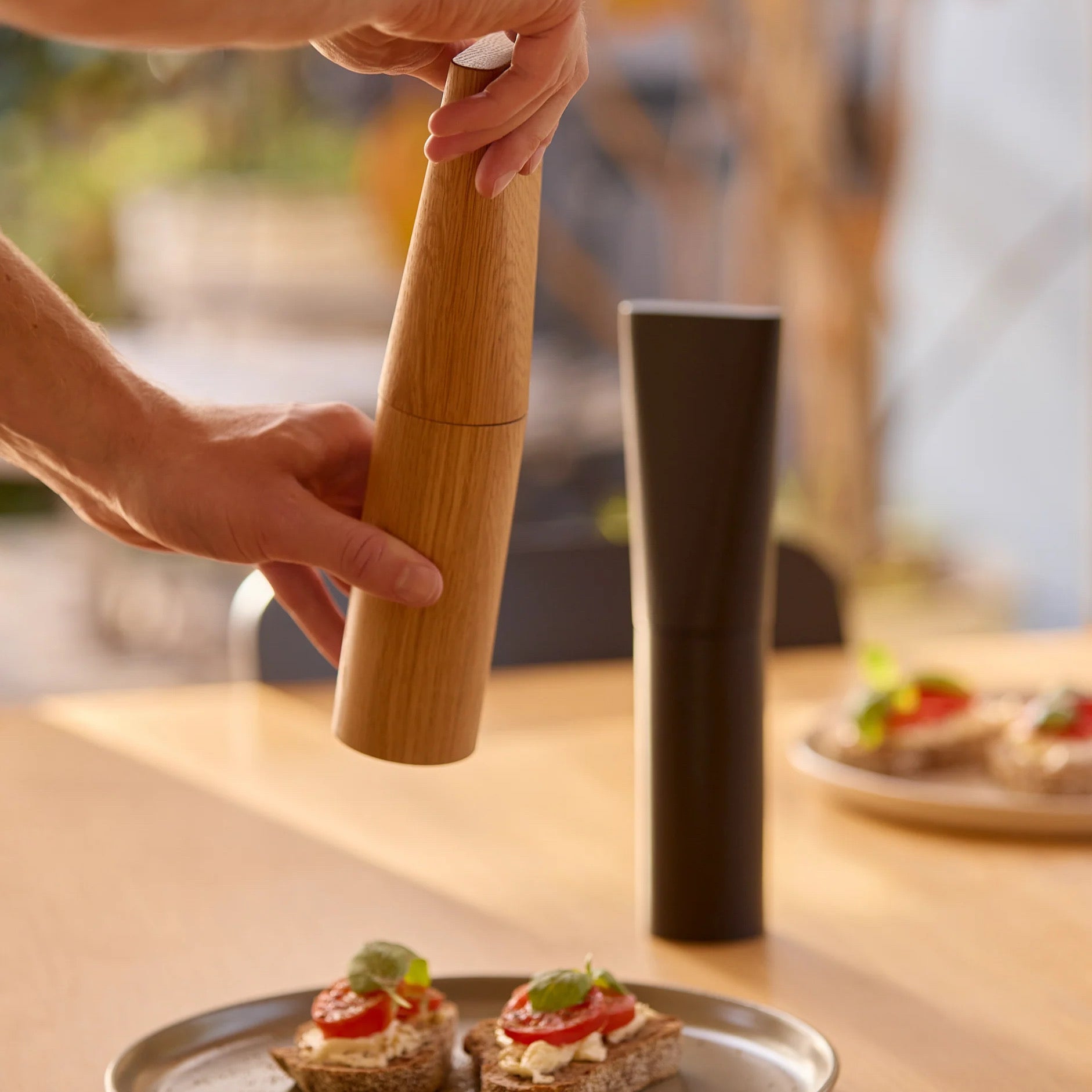 A person's hands are shown using a wooden pepper mill to grind pepper over small toast on a table, with a dark cylindrical container standing nearby in a warm, softly blurred kitchen setting.