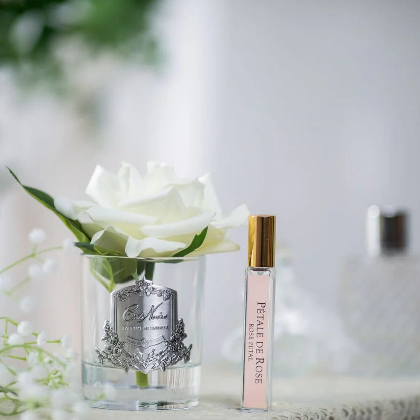 Perfume bottle labeled 'Petale de Rose' next to a white rose in a clear vase.