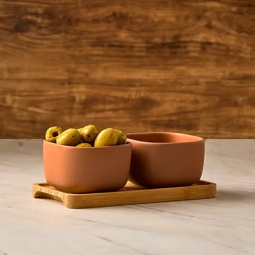 Two terracotta-colored ceramic bowls on a bamboo tray, one filled with green olives, set against a rustic wooden background and light marble surface.