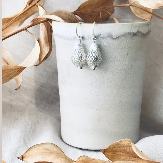 Silver earrings hanging from a white vase with dried leaves around.