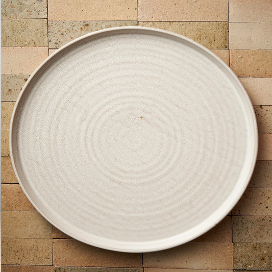 A round, white reactive glaze stoneware platter with a textured edge,