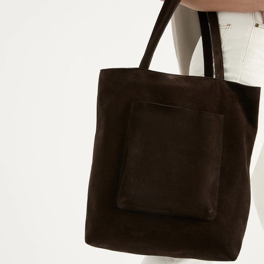 A brown suede tote bag with a large exterior patch pocket and a metal zip closure, held by a person over their shoulder.