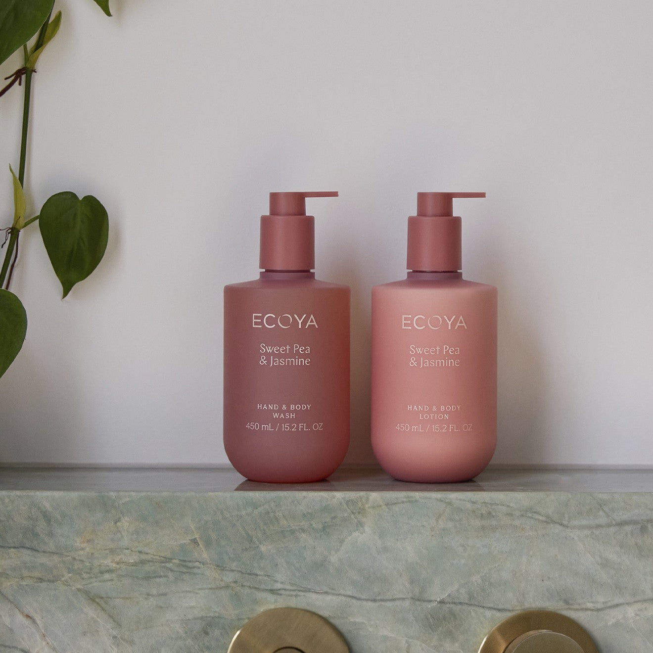 Two pink bottles of Ecoya Sweet Pea & Jasmine hand and body wash on a marble surface with a plant in the background.