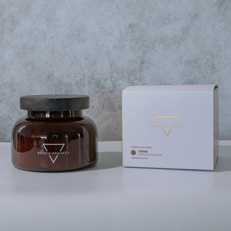A medium-sized brown jar candle with a wooden lid, alongside a white box with the 'Becca Project' logo.