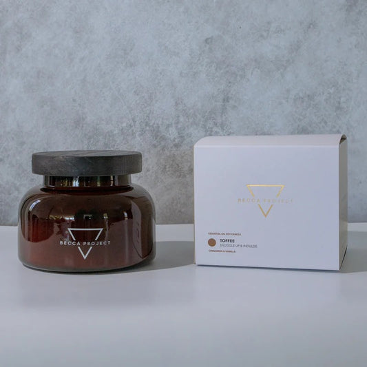 A medium-sized brown jar candle with a wooden lid, alongside a white box with the 'Becca Project' logo.