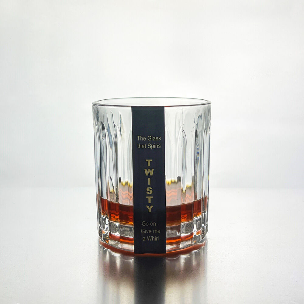 A clear glass rocks tumbler with a vertical ribbed pattern and a black paper label wrapped around the center. The label features gold text that reads 'The Glass that Spins TWISTY Give on - a Whirl'.